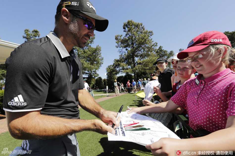 金诚在练习轮中耐心弯腰，为一位手持果岭旗的小球迷签名，周围是其他充满期待、手持纪念品的观众。