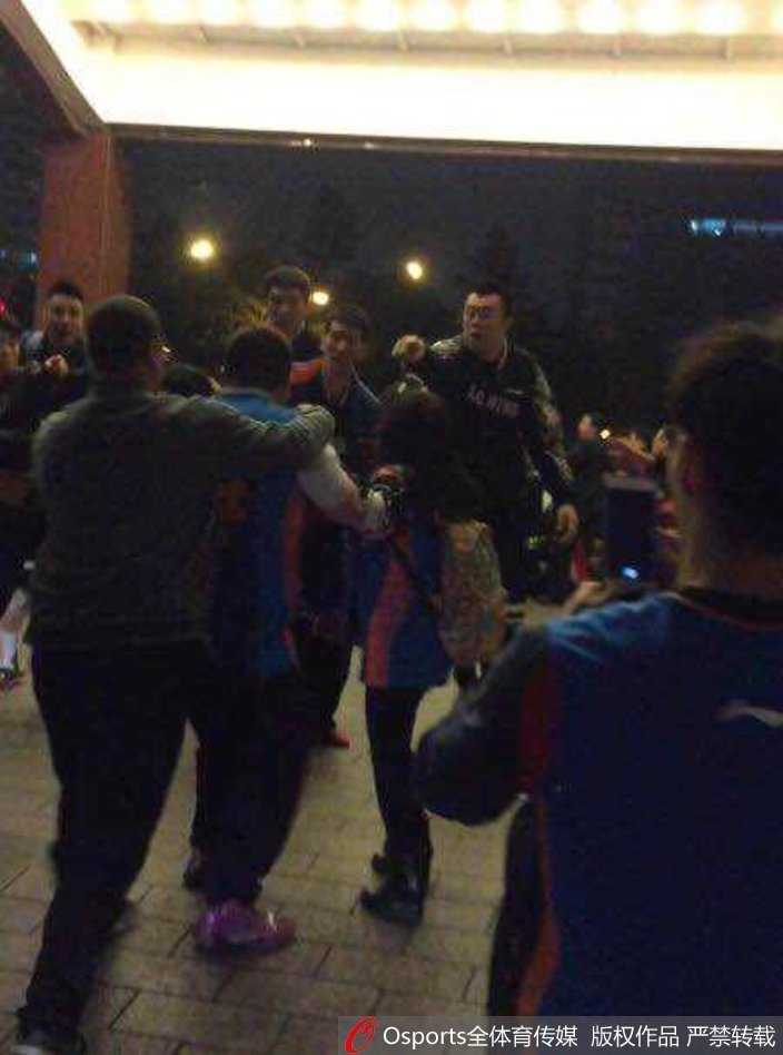 深夜的酒店门口一片混乱，多名身穿制服的人员试图挤进人群中心拉开斗殴双方，外围的球迷仍在举着手机拍摄，闪光灯与喧哗声交织。