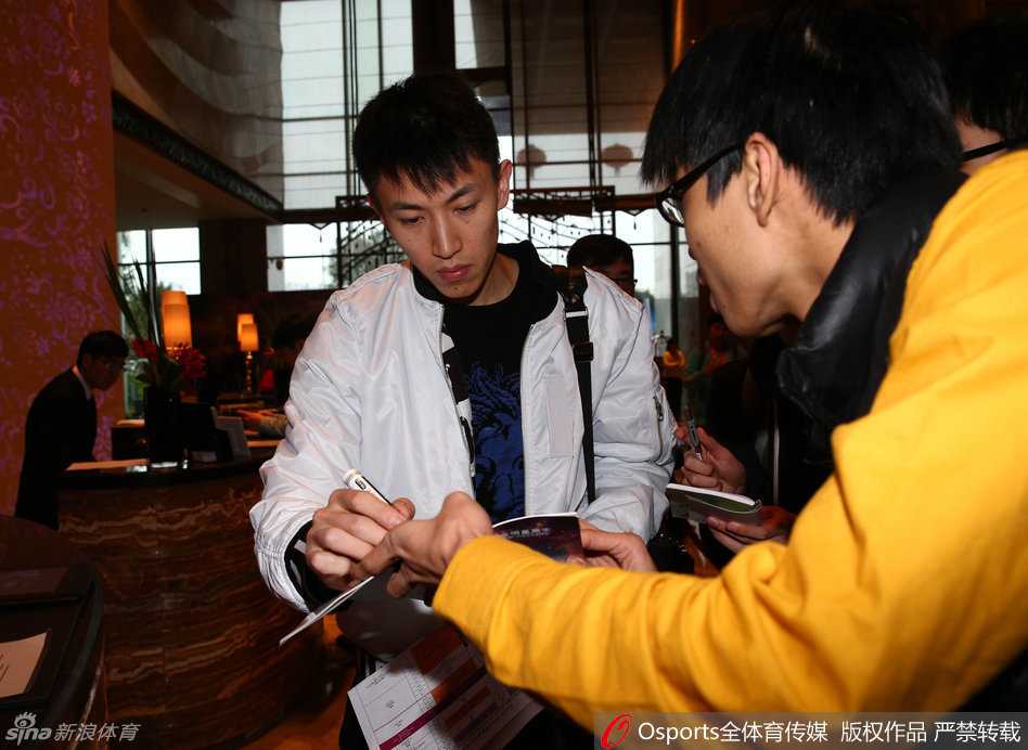 刘晓宇被几位热情球迷簇拥，他微微侧身低头，手中的笔在球衣上划过，嘴角勾起一抹宠溺的弧度，现场气氛温馨而热烈。