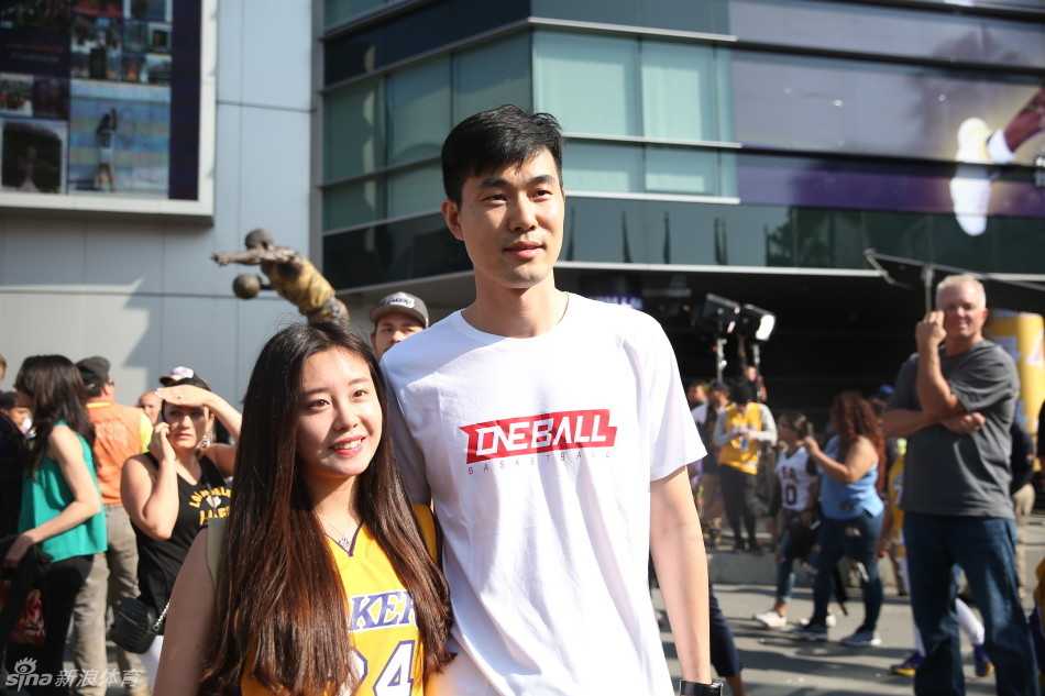 王仕鹏与女球迷合影时依然低头关注手机上的比赛信息，一旁的朱芳雨则面带微笑望向镜头，背景是人潮涌动的斯台普斯中心外场。