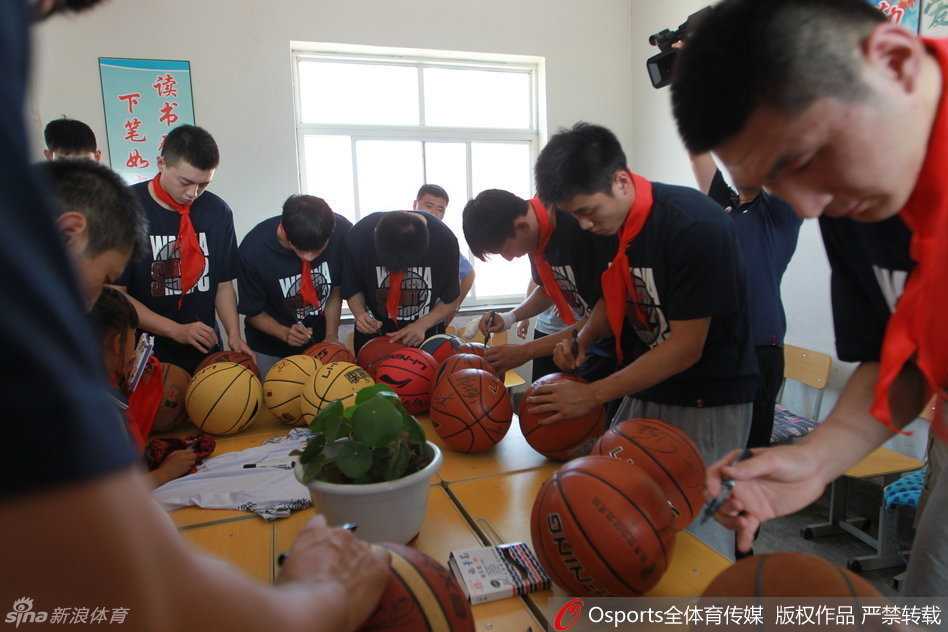 李晓旭弯腰俯身，面带微笑地为一名系着红领巾的小学生在篮球上签名，周围聚满了充满期待眼神的孩子们。