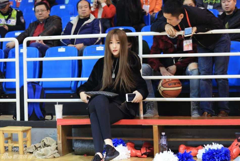 一位戴着眼镜的八一队铁杆女球迷静静坐在场边观战，她面容清秀，眼神专注地望向赛场，温柔的气质与场上激烈的对抗形成鲜明对比。