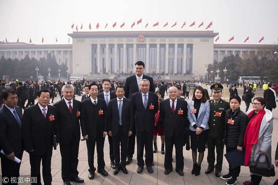 姚明在会场的走廊上与另一位委员并肩行走，他侧耳倾听对方讲话，虽然周围人潮涌动，但人们都自觉地为这个高大的身影留出一点空间，画面充满了动态的和谐感。