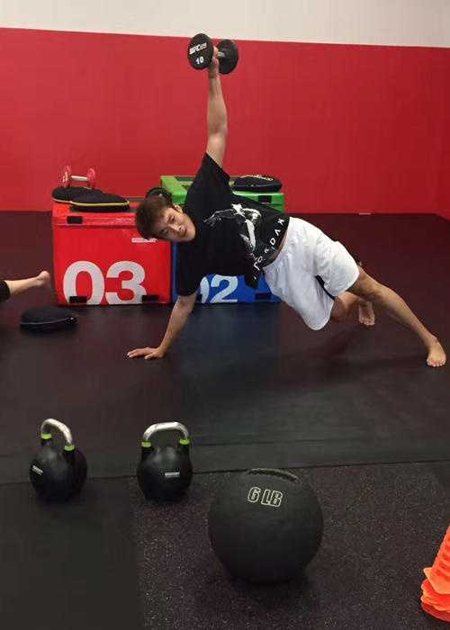 丁彦雨航身披达拉斯小牛队深蓝色训练服，在训练馆里神情专注地进行拉伸，阳光透过场馆高窗洒在他坚毅的侧脸上