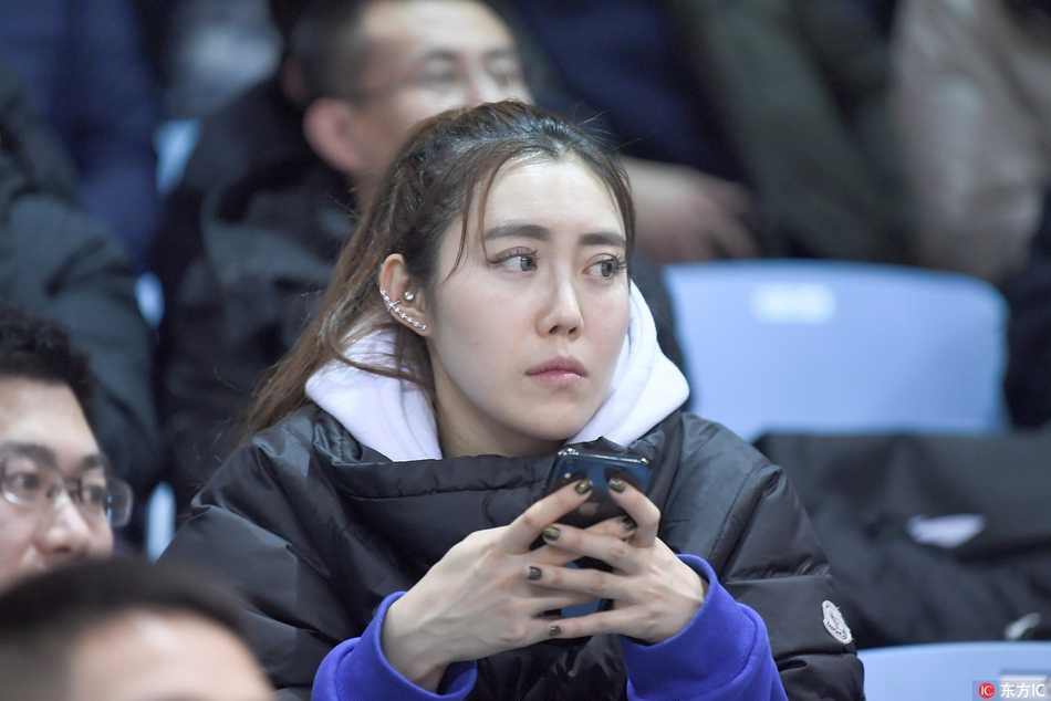 两名女生凑在一起观看手机屏幕，其中一人身穿灰色大衣，另一人戴着黑框眼镜，两人可能是中场休息时在查看心爱球员的数据，神态专注而亲密