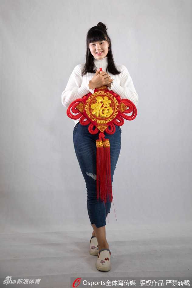 黄思静手持一张精致的金色福字卡，对着镜头俏皮地眨眼，展现出赛场之外活泼可爱的一面，背景中隐约可见其他队友正在相互整理着装，气氛温馨和谐。