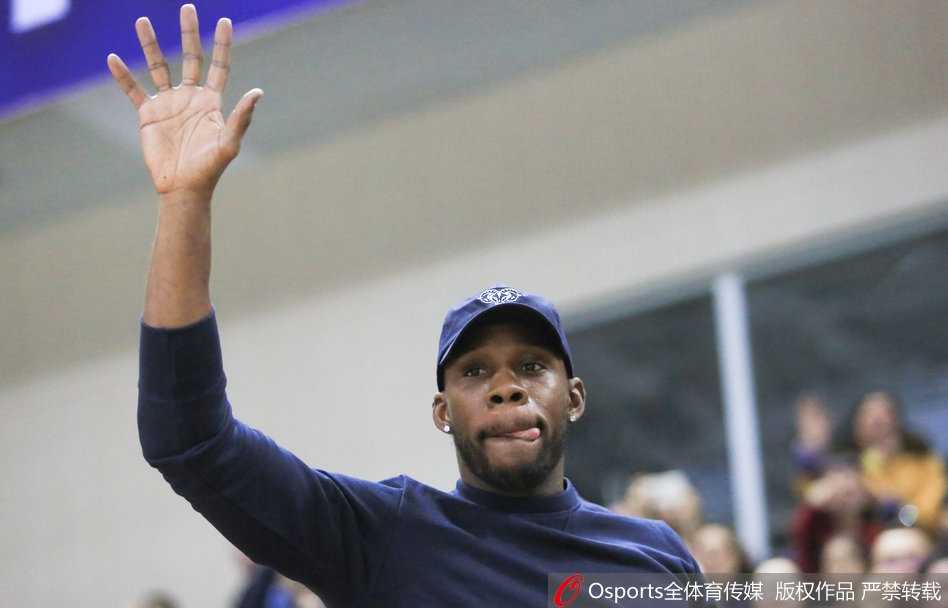 哈德森与身旁的友人交谈，脸上带着轻松的笑容，背景是电子记分牌上辽宁队领先的比分，现场热烈的氛围感染了这位篮球明星。