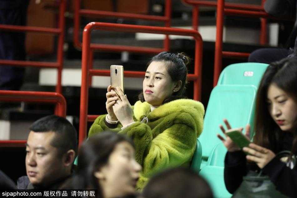 一位年轻球迷将貂绒大衣搭在肩上，内搭辽宁队的红色球衣，他面带笑容指着球场方向，仿佛在向同伴夸赞今晚主队的出色表现。