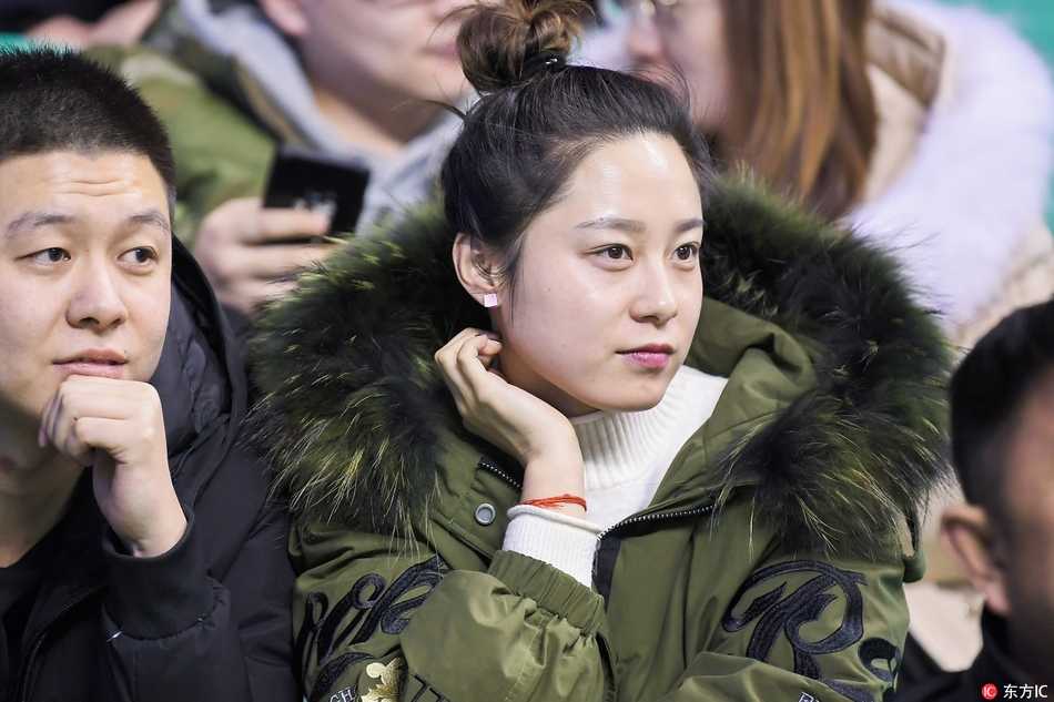 一位身着时尚白色外套的冷艳美女，安静地坐在看台上，身后是沸腾的人群，她的眼神中带着一丝对比赛胜负的淡然，更享受这冬日里的篮球之夜。