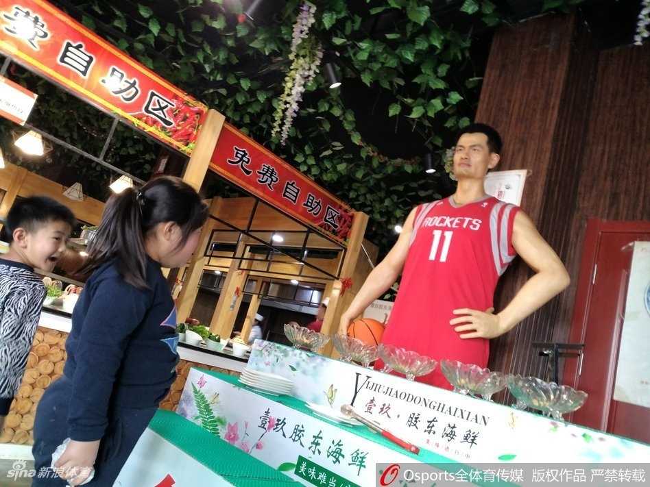 一位身形巨大的‘篮球巨星’蜡像，身穿红色火箭队战袍，安静地站在饭店大厅中央，周围食客纷纷侧目，仿佛在仰望真正的巨人
