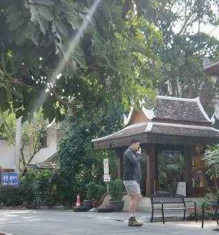 姚明一家在游览间隙停下脚步休息。姚明双手插兜，面带微笑地看向镜头，身后的绿植和泰式建筑衬托出度假的轻松氛围，尽管穿着朴素，但他那挺拔的身姿和亲和的笑容依然是画面中绝对的中心。