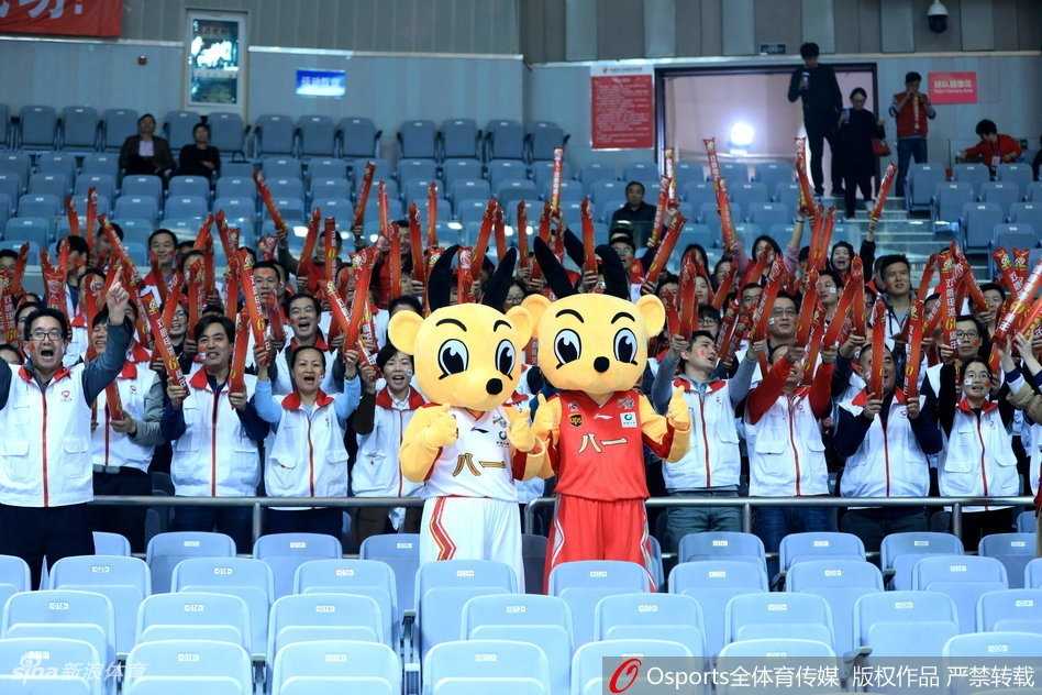 吉祥物俯身逗弄看台上的小朋友，它用毛茸茸的手掌轻轻抚摸孩子的头，周围的家长纷纷举起手机记录下这暖心一刻，整个看台区域洋溢着欢乐的气氛