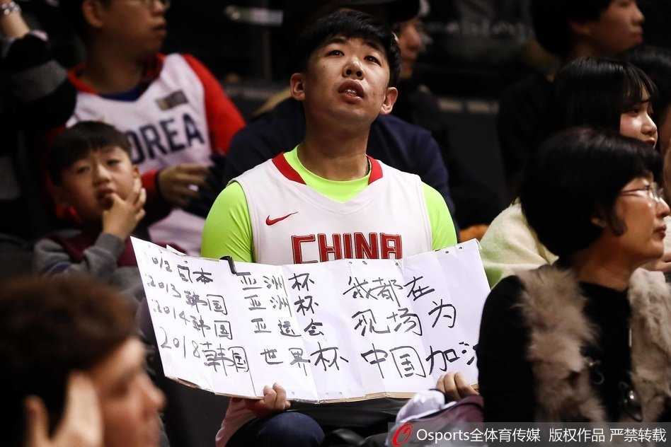 这位戴着可爱发卡的女生正对着镜头比出胜利的手势，她的笑容如同冬日里的暖阳，灿烂而治愈，手中的加油牌在灯光下熠熠生辉，仿佛整个赛场都被她的热情所感染。