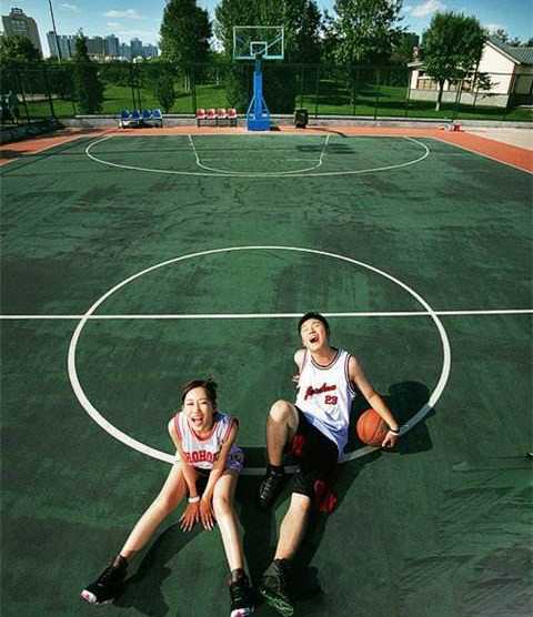 俯拍视角下，新人在巨大的篮球场中线上相对而坐，新娘的婚纱裙摆如花瓣般散开铺陈在地板上，他们正用粉笔写下属于彼此的爱情誓言。