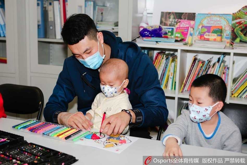 翟晓川盘腿坐在地板上，与几个戴着口罩的小朋友围成一圈，他正拿着一个彩色气球逗其中一个孩子，孩子们咯咯笑着伸手去够，场面温馨如同一个大哥哥带着弟弟妹妹做游戏