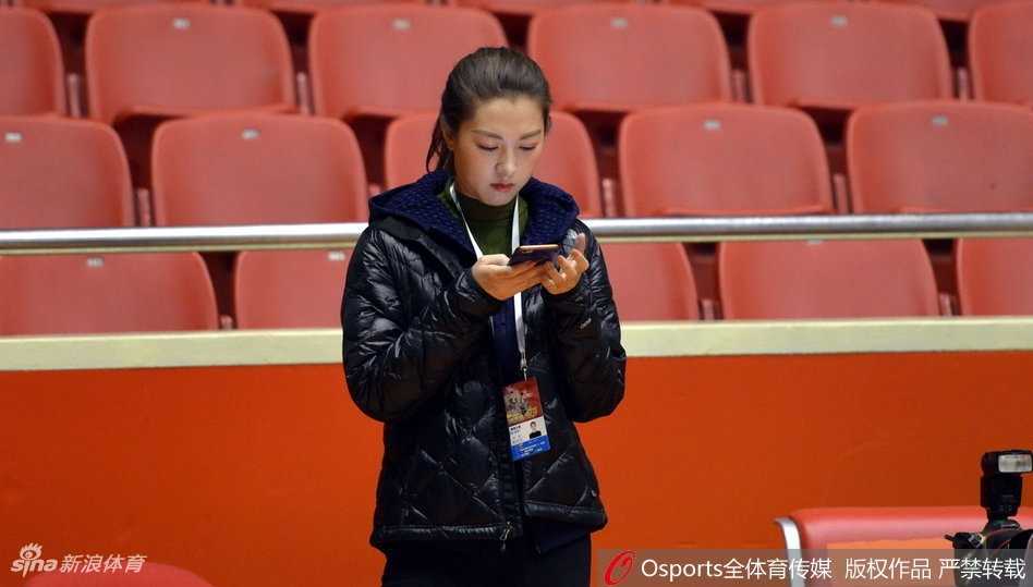 在明亮的训练场馆中，新疆男篮队员们正围绕教练认真聆听战术板上的布置，场边的美女记者手持笔记本，眼神专注地观察着球队的每一次合练细节，画面充满了备战的紧张感。