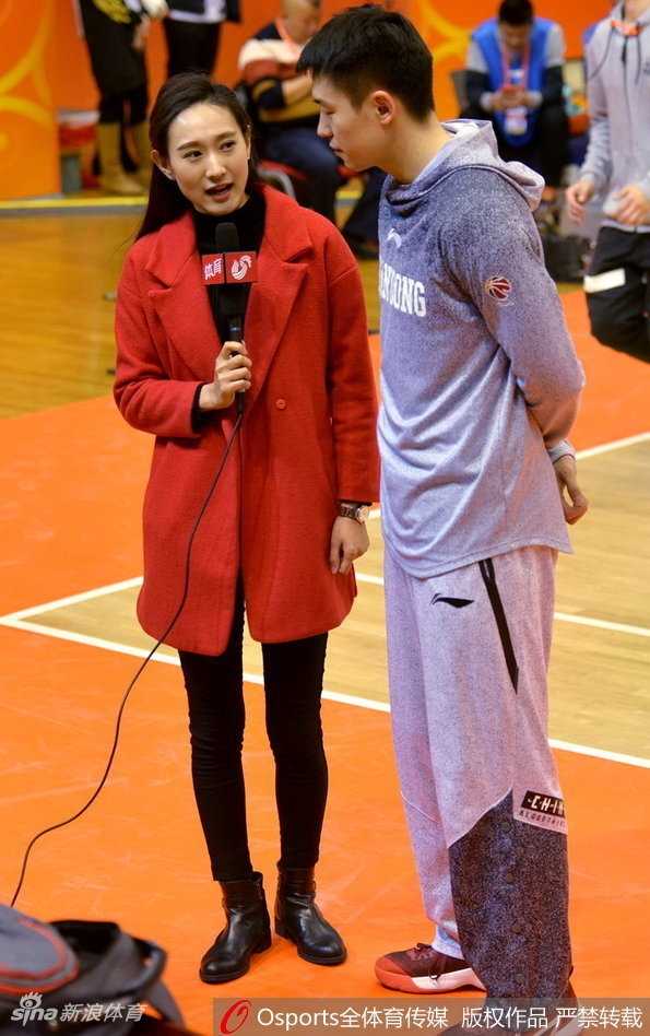 一位美女记者身着简约的灰色大衣，在球场通道内回眸一笑，她颈间挂着醒目的媒体证件，背景是略显模糊的球员通道，动静结合之间，仿佛凝固了赛前那一刻的紧张与期待。